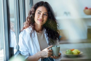 Güzel bir kadının bir fincan kahve içerken evinin penceresinin yanında dikilirken çekilmiş fotoğrafı..