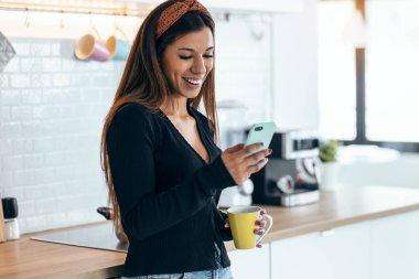 Shot of beautiful woman using her mobile phone while drinking a cup of coffee in the kitchen at home.