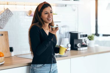 Shot of beautiful woman using her mobile phone while drinking a cup of coffee in the kitchen at home.