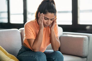 Evdeki oturma odasında kanepede otururken sorunları düşünen endişeli genç bir kadın..