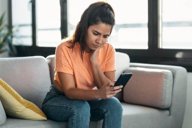 Evdeki oturma odasındaki kanepede oturan sorunlarını düşünürken endişeli genç bir kadının cep telefonunu kullanması..