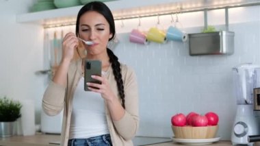 Video of pretty woman sending messages with mobile phone while eating yogurt int he kitchen at home.