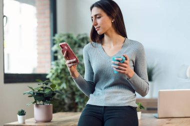 Zeki bir iş kadınının evdeki oturma odasında masaya yaslanmış bir fincan kahve içerken cep telefonu kullanması..