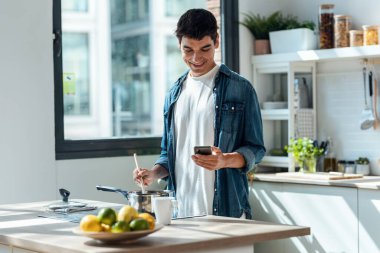 Shot of handsome young man cooking and using his mobile phone in the kitchen at home.