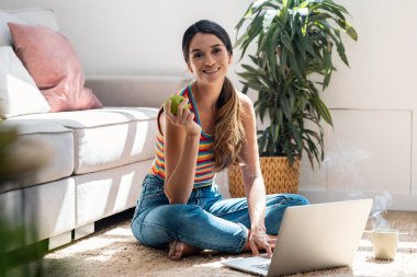 Shot of pretty young woman using her laptop while eating an apple sitting on the floor at home.