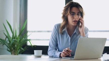 Ofiste laptopuyla çalışırken cep telefonuyla konuşan güzel bir iş kadınının videosu.