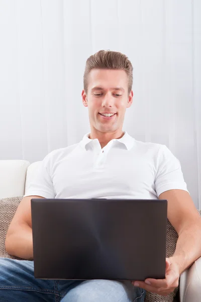 Gelukkig jonge man zittend op de Bank met behulp van laptop — Stockfoto