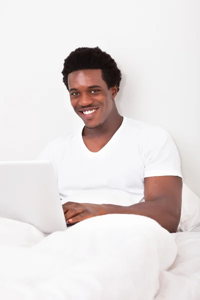 Χαρούμενος νεαρός χρησιμοποιώντας φορητό υπολογιστή στο κρεβάτι — Φωτογραφία Αρχείου