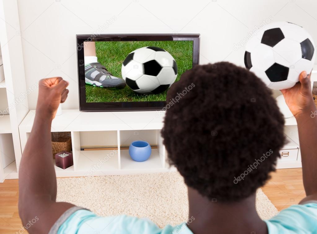 African Young Man Watching Television Holding Football