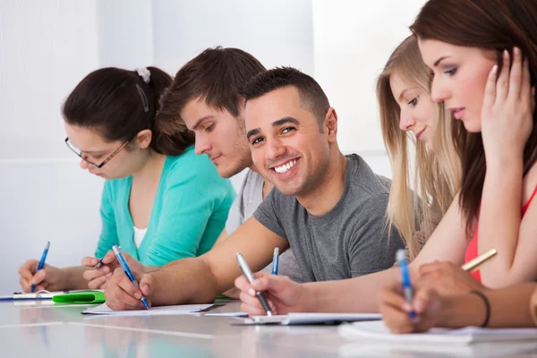 Knappe student zitten met klasgenoten schrijven aan balie — Stockfoto