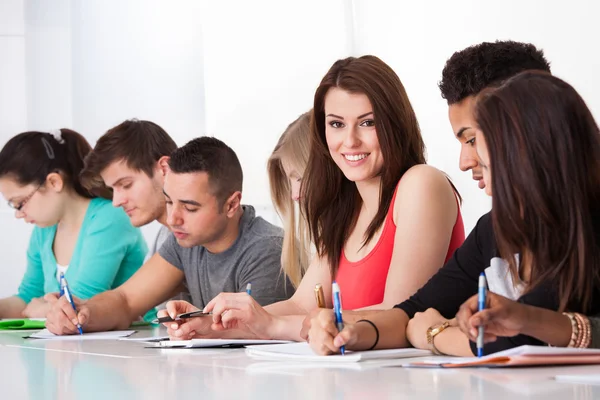 Mooie student zitten met klasgenoten schrijven aan balie — Stockfoto