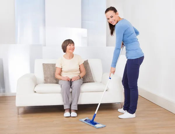 Cuidador de limpeza chão enquanto mulher sentado no sofá — Fotografia de Stock