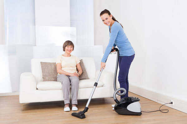 Caretaker Cleaning Floor While Senior Woman Sitting On Sofa