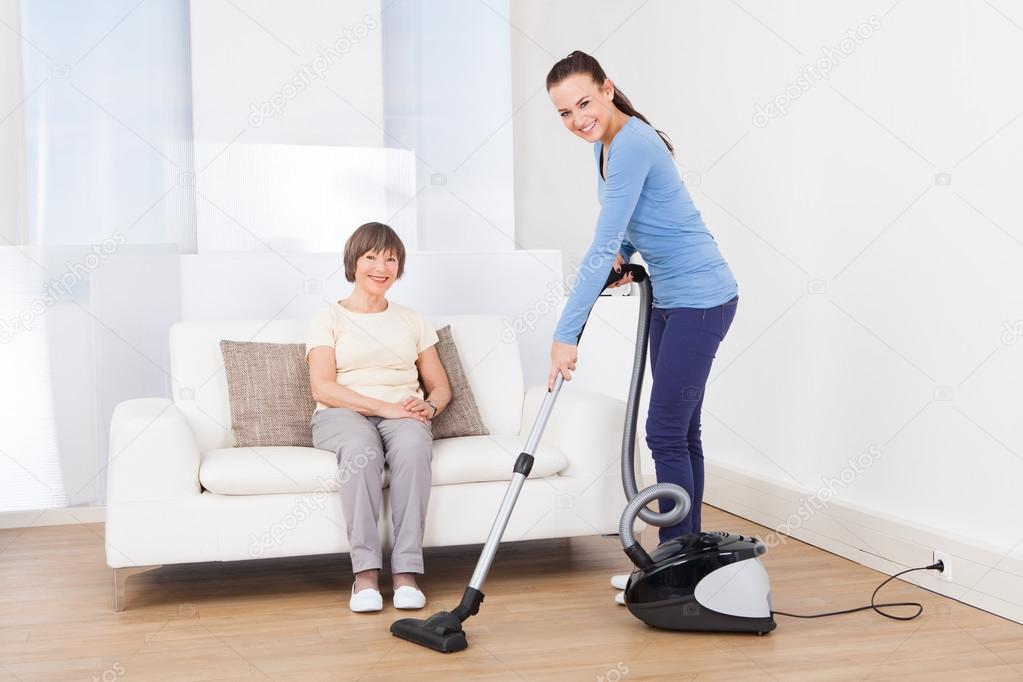 Caretaker Cleaning Floor While Senior Woman Sitting On Sofa — Stock