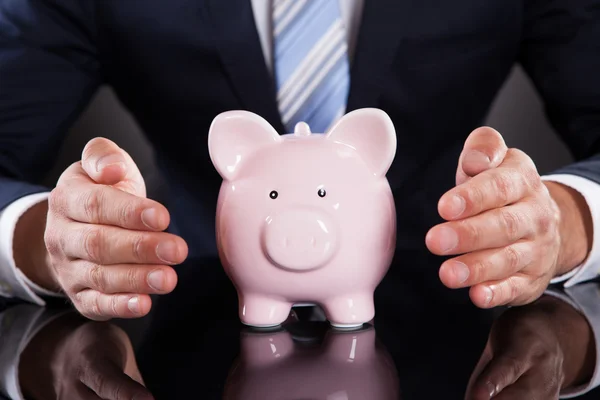 Empresario protegiendo Piggybank con las manos en el escritorio — Foto de Stock