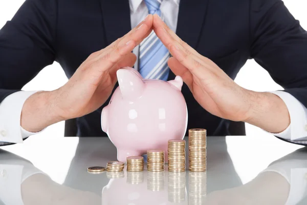 Empresário abrigando moedas e Piggybank na mesa — Fotografia de Stock