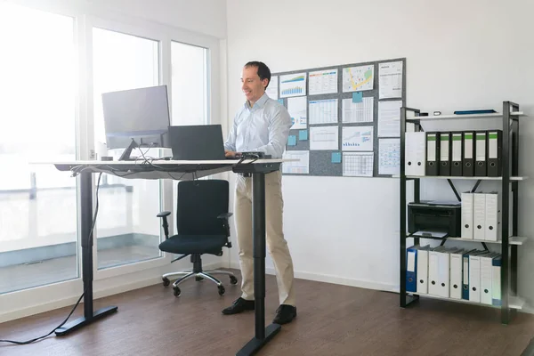 良い姿勢のためにオフィスで調節可能な高さ立ちデスクを使用して男 — ストック写真