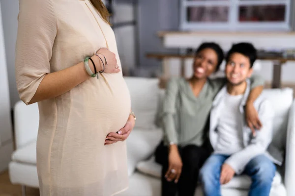 怀孕的女人摸着她心爱的女人站在微笑的年轻夫妇的面前 — 图库照片