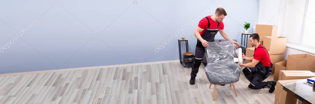 Two Young Male Movers Packing Furniture With Plastic Wrap In Living Room