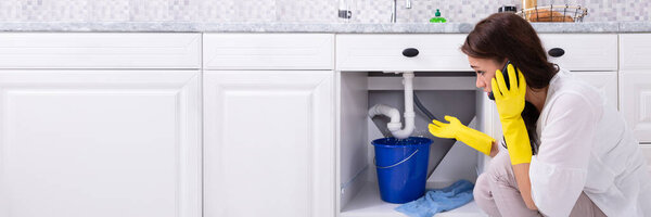 Sad Young Woman Calling Plumber In Front Of Water Leaking From Sink Pipe