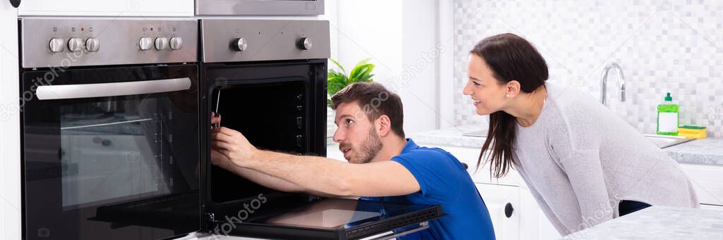 Smiling Young Woman Looking At Technician Fixing Oven In Kitchen