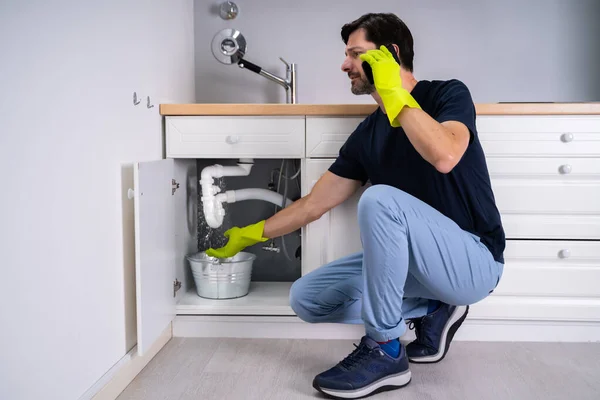 Sad Young Man Calling Plumber In Front Of Water Leaking From Sink Pipe