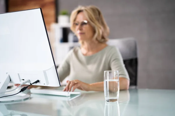 Water Drinking Glass On Desk And Woman In Foreground Using Computer