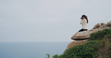 Kadın dağın tepesinde oturur ve fotoğraf çekmek için kamera kullanır.