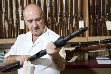 Mature gun shop merchant looking at rifle in store clipart