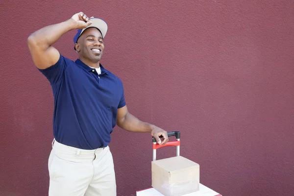 Happy young African American delivery man standing with handtruck over colored background — Stock Photo, Image