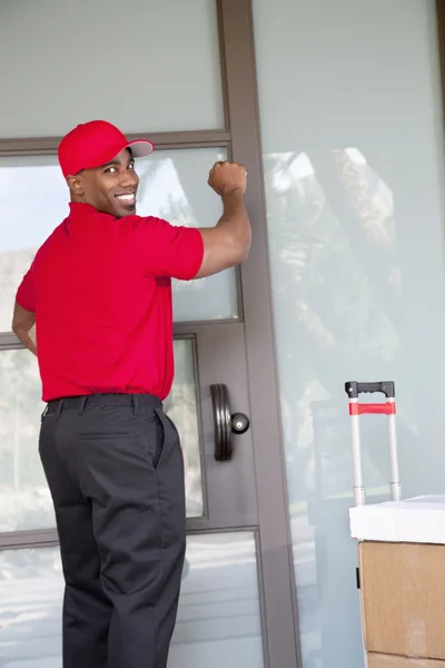Jovem entregador com pacotes olhando para trás enquanto bate na porta — Fotografia de Stock