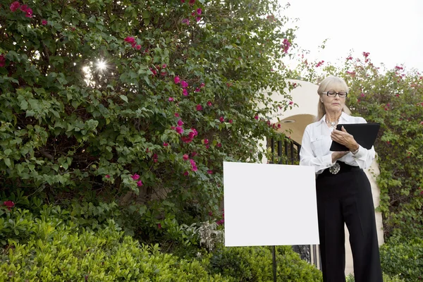 Senior real estate agent standing by sign board reading from clipboard — Stock Photo, Image