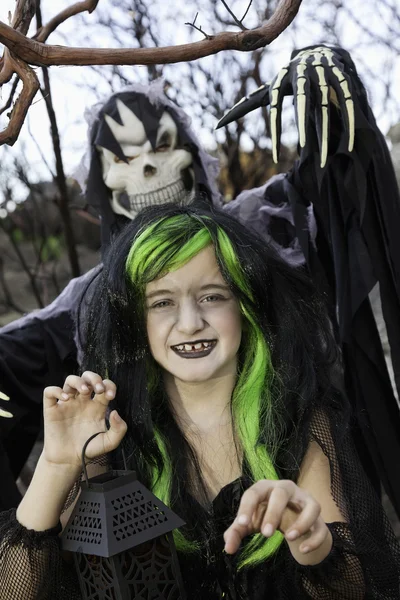 Little witch holding lantern with person costumed as grim reaper standing in background — Stock Photo, Image