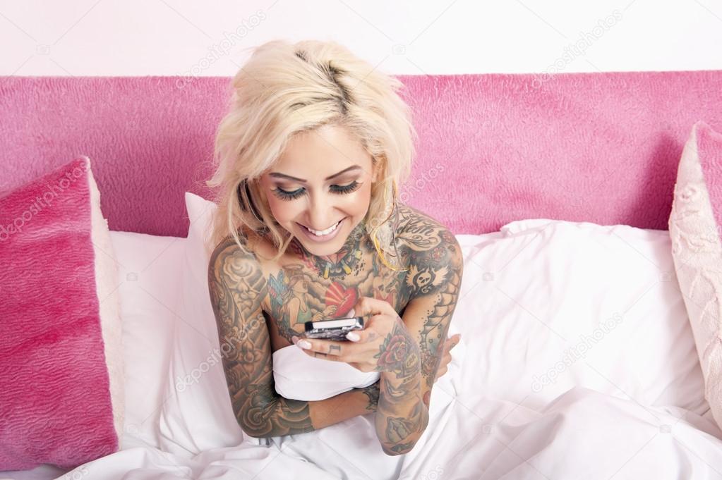 Naked woman with tattoo sitting in bed reading text message in mobile phone