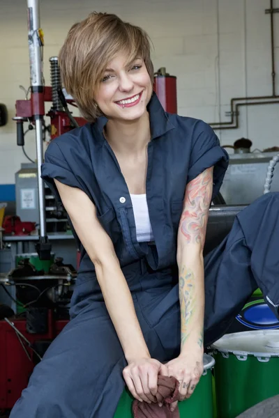 Retrato de una joven mecánica feliz sentada sobre un tambor de aceite en un taller de automóviles —  Fotos de Stock