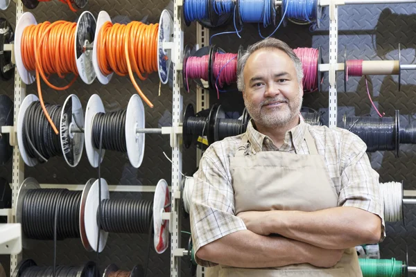 Portrait of a happy mature salesperson standing in front of electrical wire spool with arms crossed in hardware store — Stock Photo, Image