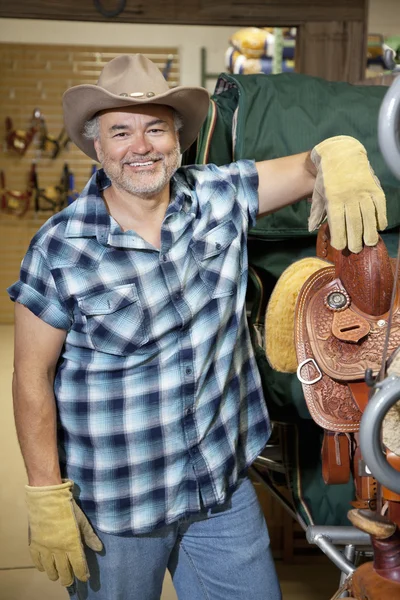 Retrato de um feliz maduro cowboy de pé por sela no feed store — Fotografia de Stock