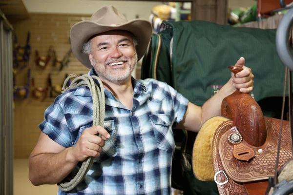 Happy mature cowboy with saddle and rope in feed store — Stock Photo, Image