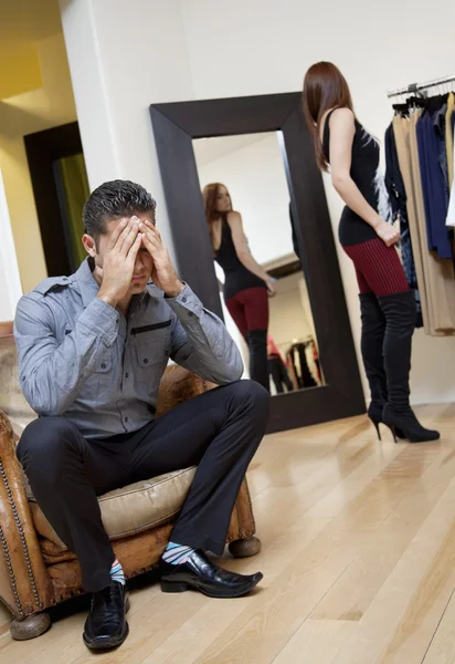 Worried young man sitting on armchair with woman in background trying on clothes — Stok fotoğraf