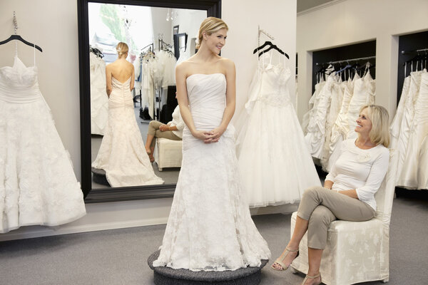 Happy mother looking at young daughter dressed in wedding gown in bridal boutique