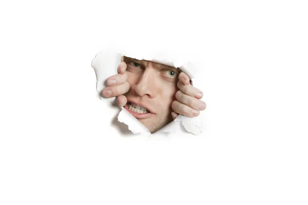 Portrait of a aggressive mid adult man peeking from ripped white paper hole — Stock Photo, Image