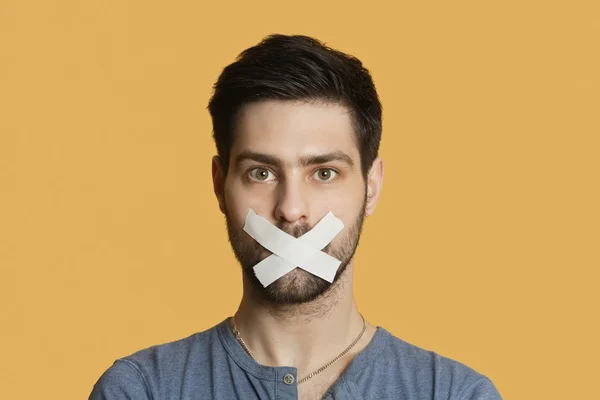 Portrait of a young man with tape on mouth over colored background — Stock Photo, Image