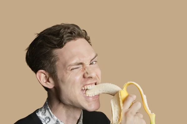 Portrait of a mid adult man winking while biting banana over colored background — Stock Photo, Image