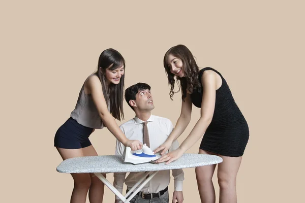 Шокированный мужчина смотрит на молодых женщин гладильная галстук на цветном фоне — стоковое фото