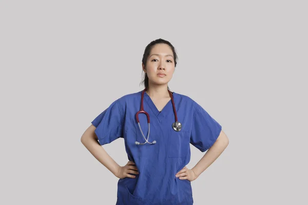 Portrait of a confident young Asian female doctor standing with hands on hips over gray background — Stock Photo, Image