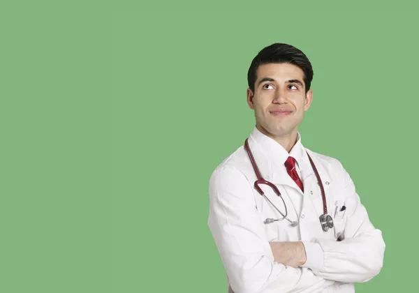 Médico varón con bata de laboratorio de pie con los brazos cruzados mirando hacia arriba sobre fondo verde —  Fotos de Stock
