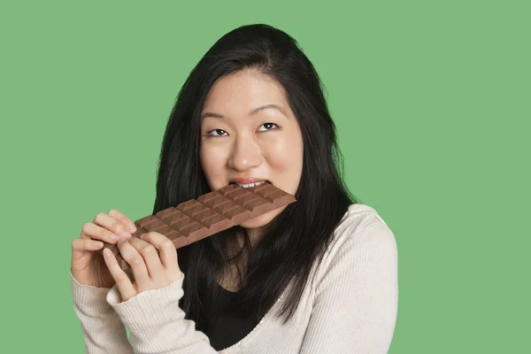 Cute young woman eating a large chocolate bar over green background — Stock Photo, Image