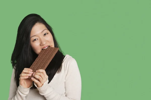 Porträt einer jungen Frau, die einen großen Schokoriegel vor grünem Hintergrund isst — Stockfoto