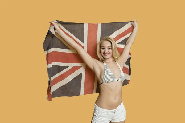 Portrait of a beautiful young woman holding British flag with arms raised over colored background Royalty Free Stock Photos