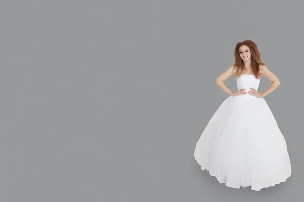 Portrét šťastné brunetka ve svatebních šatech s rukama na bocích stojí nad šedým pozadím — Stock fotografie